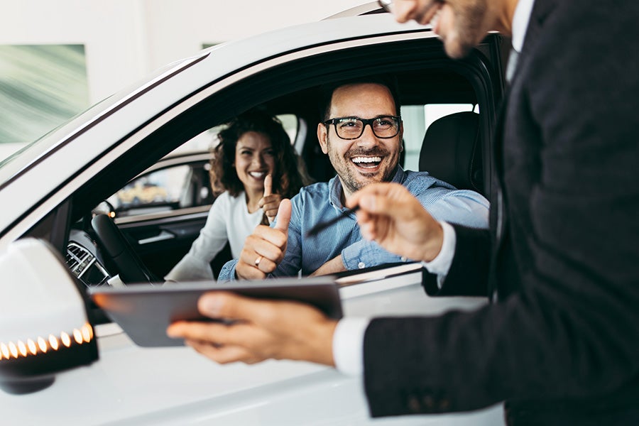 A man and women sitting inside car are smiling and showing thumbs up to salesperson.